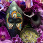 May include: Close-up view of a dark-colored, beaded African mask adorned with cowrie shells and vibrant orange, green, and blue beads. The mask is nestled amongst a lush arrangement of deep purple, magenta, and lavender flowers, including orchids and calla lilies. A small, shallow bowl filled with dried, light green herbs with hints of purple sits in front of the mask.