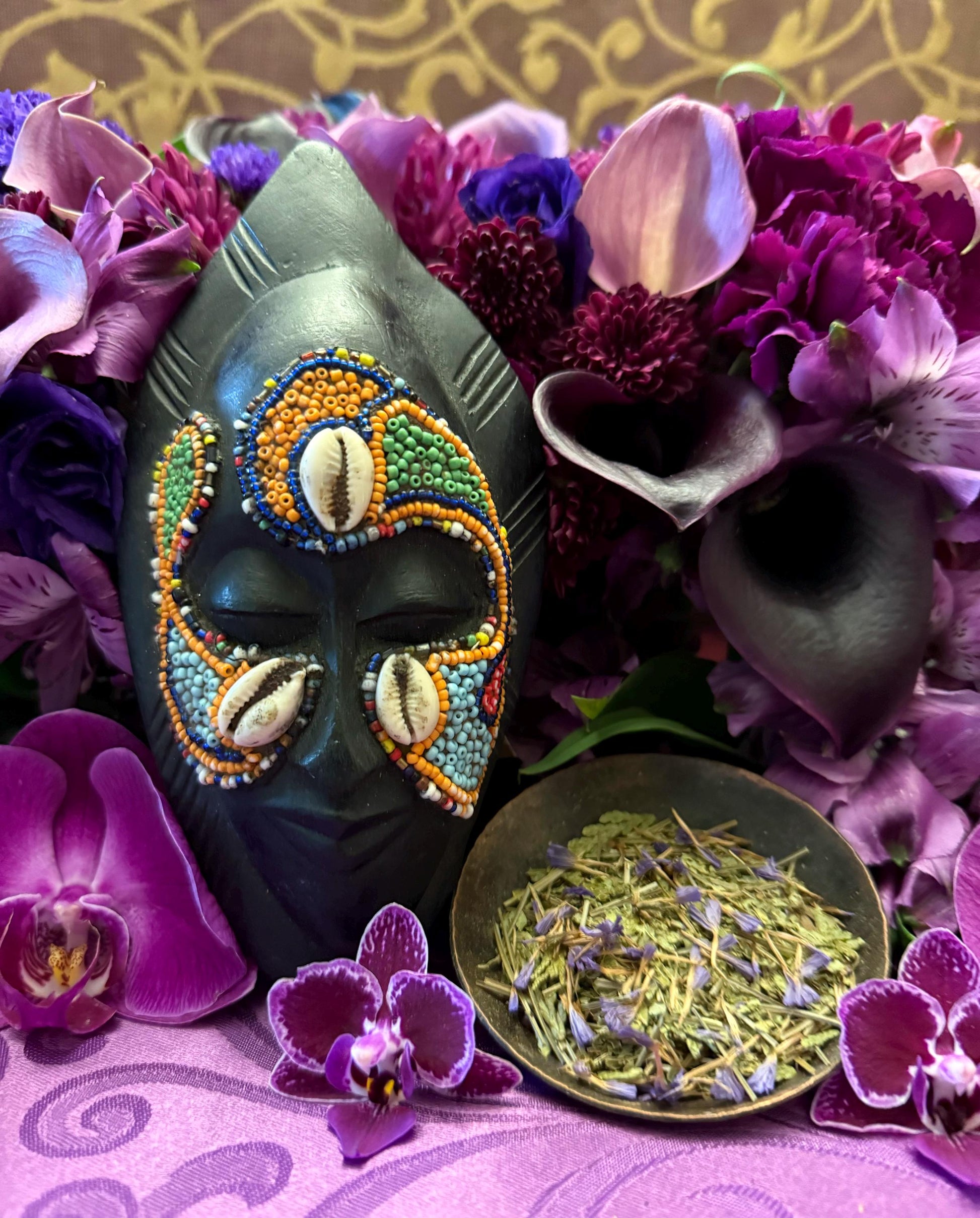 May include: Close-up view of a dark-colored, beaded African mask adorned with cowrie shells and vibrant orange, green, and blue beads. The mask is nestled amongst a lush arrangement of deep purple, magenta, and lavender flowers, including orchids and calla lilies. A small, shallow bowl filled with dried, light green herbs with hints of purple sits in front of the mask.